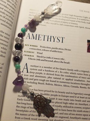 A beaded bracelet of amethyst, clear quartz and green beads with a silver hamsa charm, resting on an open book page headed Amethyst.