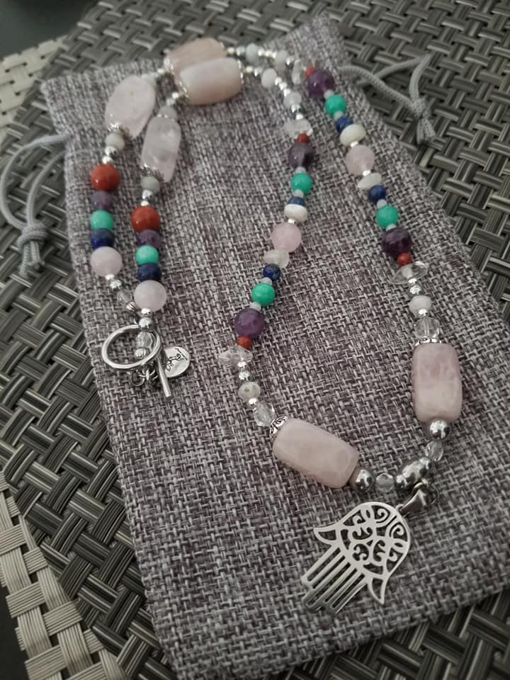 Two beaded necklaces combining rose quartz, amethyst, coral and turquoise beads with a silver hamsa pendant, laid on a grey drawstring pouch.