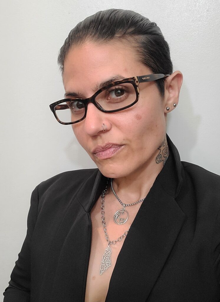 Ren Morales in a black blazer and tortoiseshell glasses, wearing two silver chain necklaces with a swirl pendant and a filigree drop.