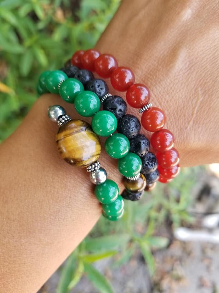 Three stacked beaded bracelets in green, black lava stone with tiger’s eye, and red, worn on a wrist outdoors.