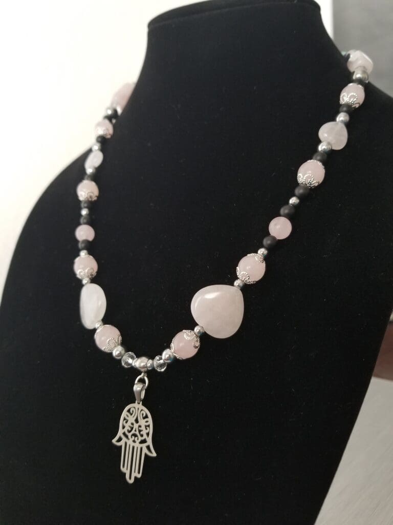 A rose quartz and black bead necklace with silver filigree caps and a silver hamsa hand pendant, on a black velvet bust.