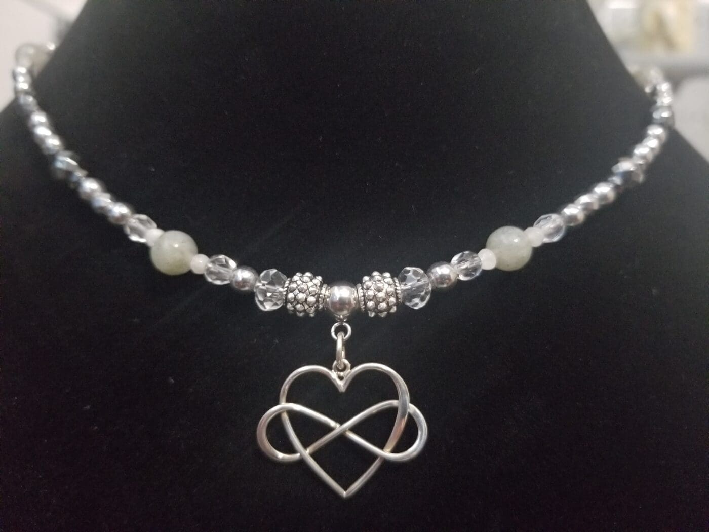 A silver and clear crystal beaded necklace with pale green jade beads and an interlocking heart and infinity pendant, on a black velvet bust.