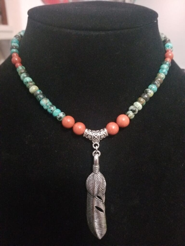 A necklace of turquoise, African turquoise and coral-coloured beads with a large silver feather pendant, on a black velvet bust.