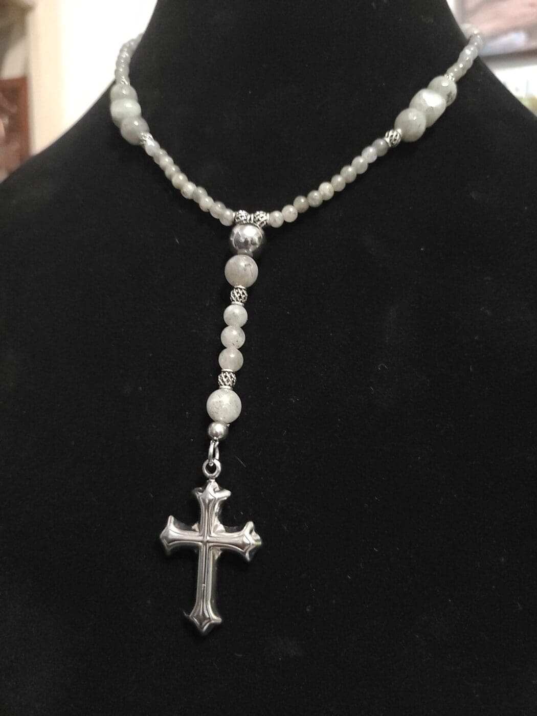 A grey labradorite beaded necklace with silver filigree spacers and a silver cross pendant, on a black velvet bust.