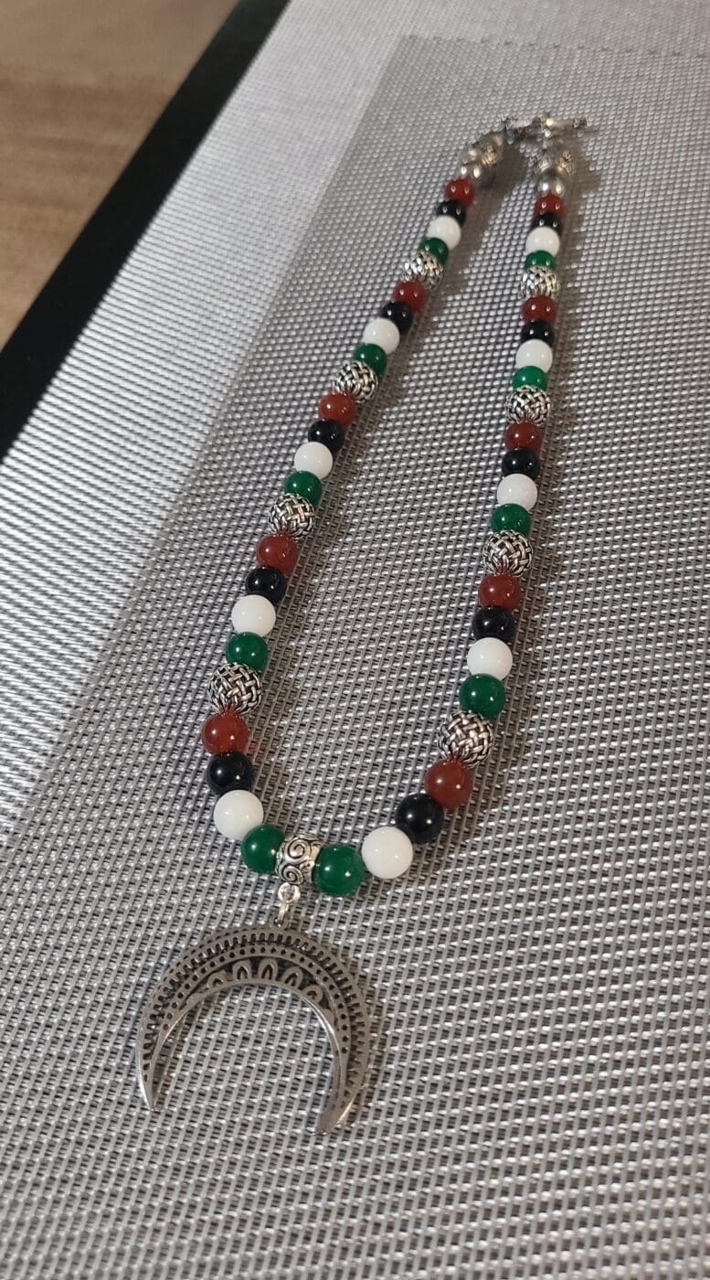 A beaded necklace of red, white, green and black stones with silver woven accent beads and a crescent horn pendant, laid on a grey woven mat.