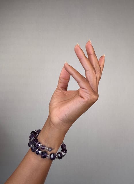 A raised hand against a grey backdrop wearing a double-wrapped bracelet of faceted purple amethyst beads with silver accents.