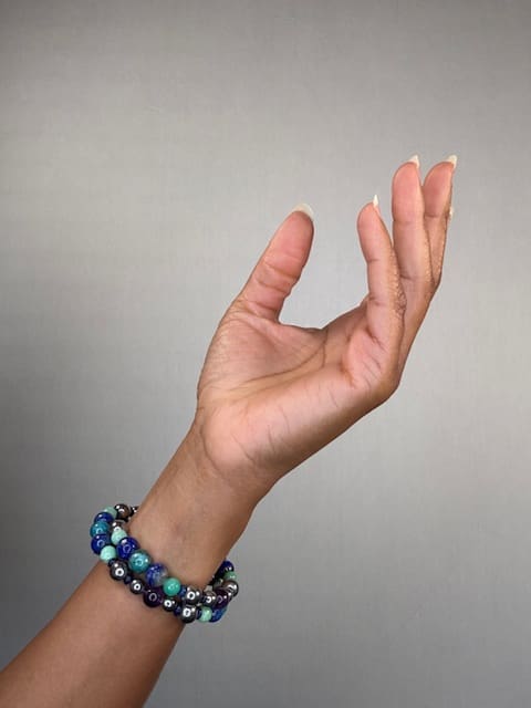 A raised hand against a grey backdrop wearing a double-wrapped bracelet of blue, teal and deep navy beads.