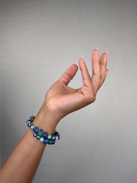 A raised hand against a grey backdrop wearing a double-wrapped bracelet of blue and turquoise beads with a patterned focal bead.