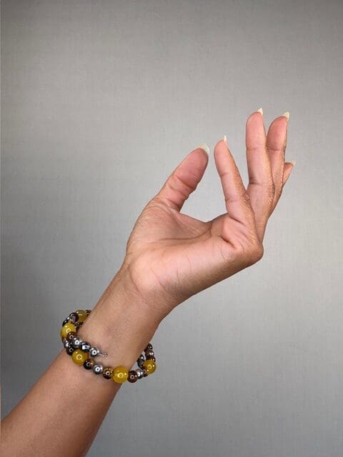 A raised hand against a grey backdrop wearing a double-wrapped bracelet of mustard yellow and brown beads with silver accents.