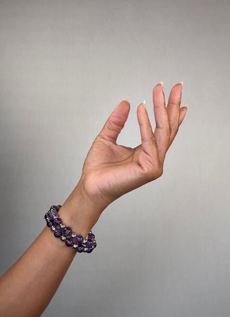 A raised hand against a grey backdrop wearing a double-wrapped bracelet of smooth purple amethyst beads with silver accents.